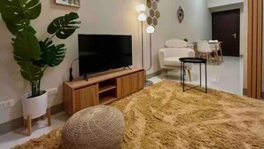 Sala de estar com televisão de tela plana sobre um suporte de madeira, uma planta grande em vaso, um tapete fofo e uma poltrona branca.