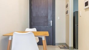 Um corredor estreito com uma porta de madeira escura no final, uma pequena mesa com duas cadeiras brancas e um frigorífico à direita.