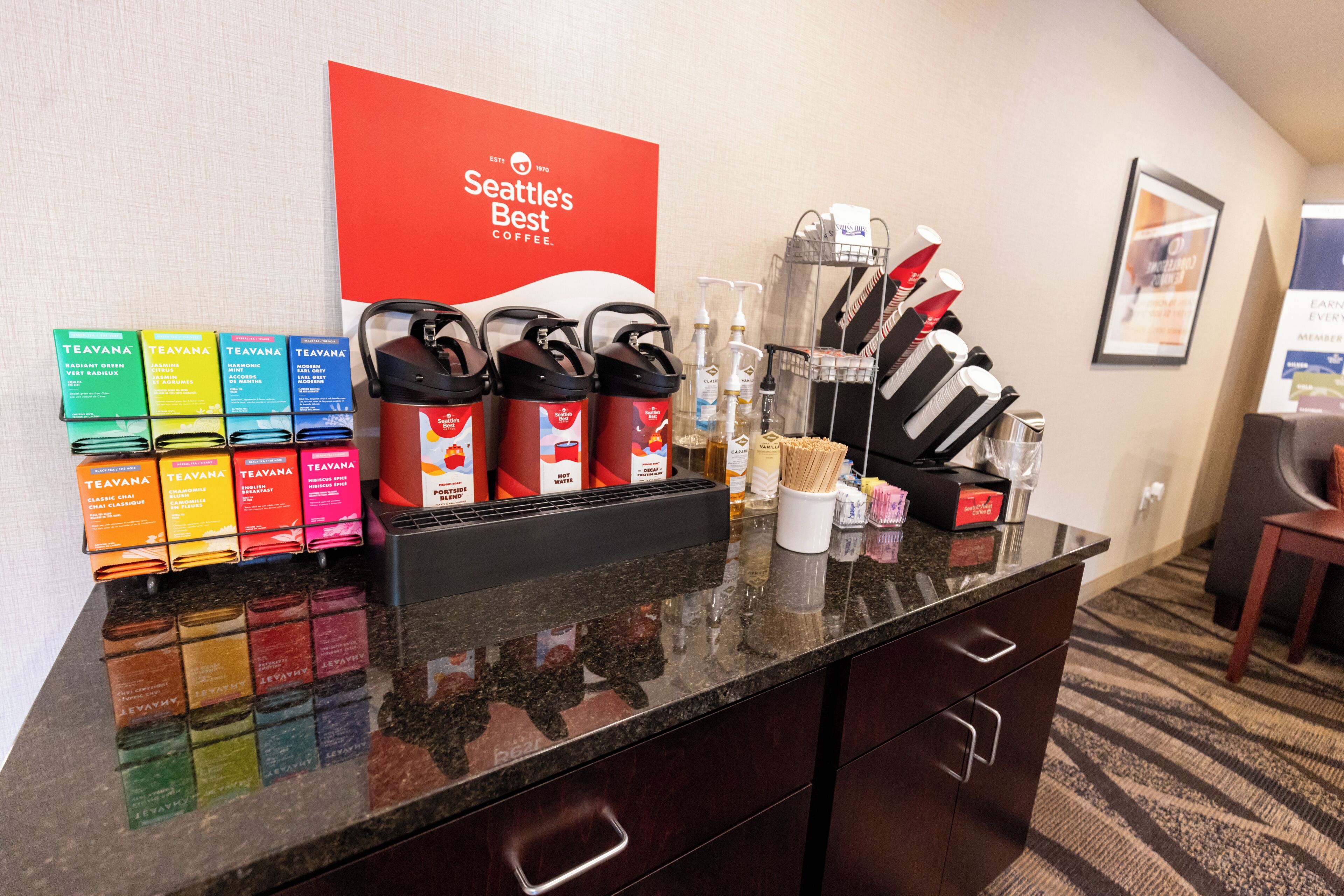 Zona de recepción de un hotel con una estación de café, un letrero rojo que dice "Seattle's Best Coffee" y varias comodidades como cajas de té, cafeteras y utensilios.