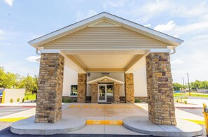 A building entrance with a covered porch supported by stone pillars, a clear sky, and a parking lot with marked parking spaces.