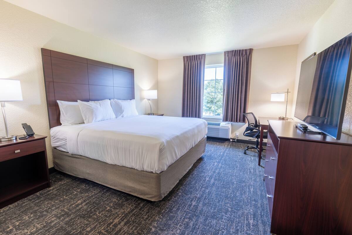 A hotel room with a large bed, a desk with a computer, a chair, a TV, and a window with curtains.