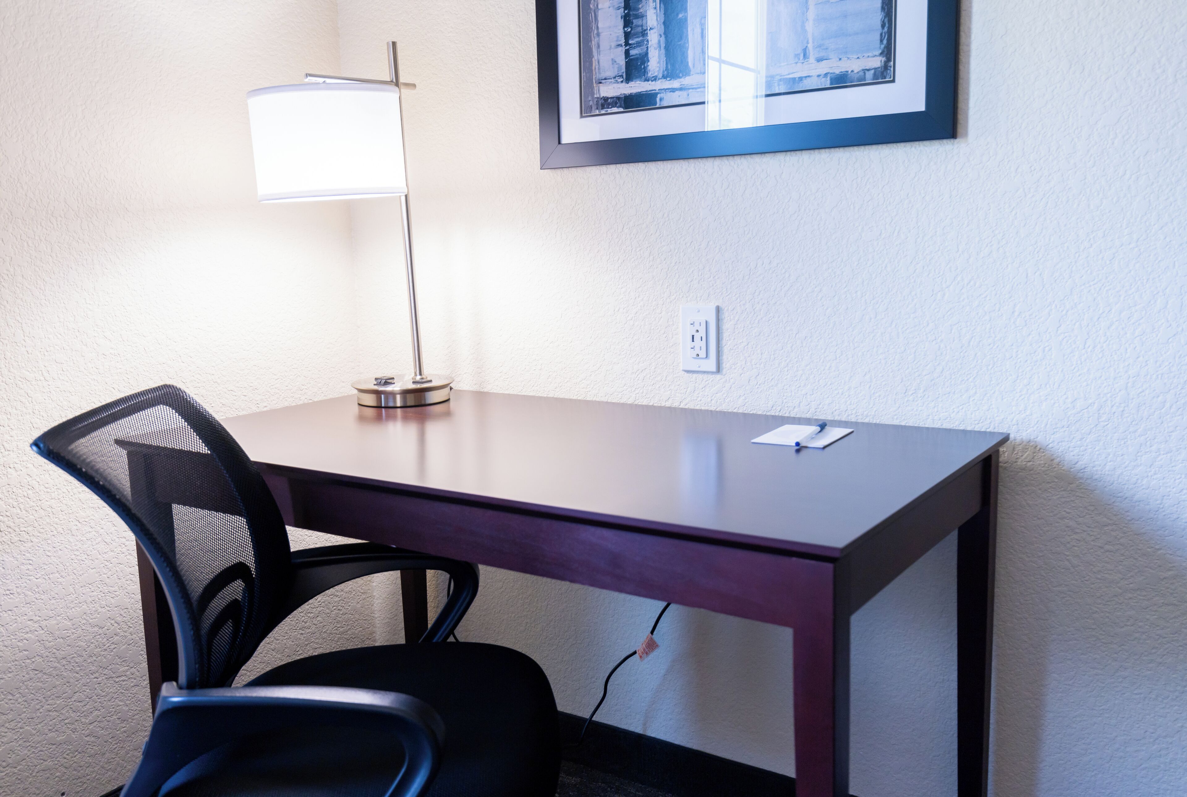 Uma mesa com cadeira, luminária e quadro na parede.