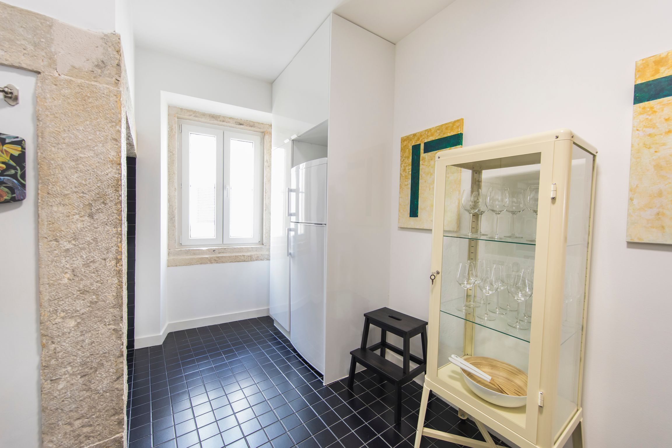 Une petite pièce étroite, avec un sol carrelé de carreaux noirs, une porte blanche, une fenêtre et une vitrine contenant des verres à vin.