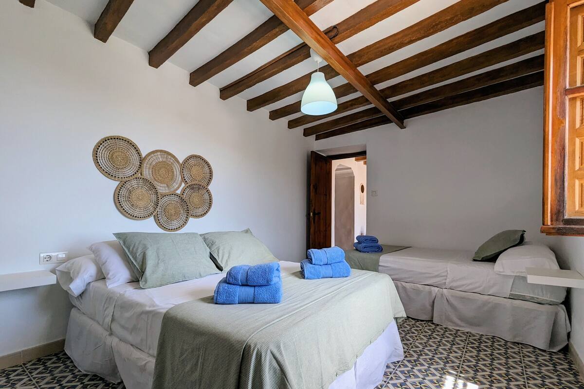A bedroom with a bed, a smaller bed, a wall-mounted light fixture, and a woven wall decoration.