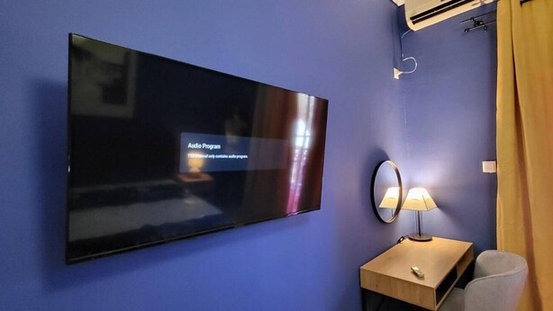 Una habitación con una televisión de pantalla plana mostrando un programa de audio, una mesita de noche con lámpara, un espejo y una silla.