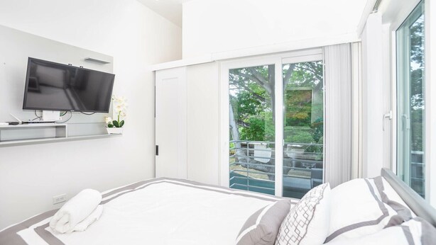 A modern hotel room with a large bed, a flat-screen TV mounted on the wall, and a sliding glass door leading to a balcony with a view of greenery. - New modern condo 3 minute walk to West Bay Beach (West Bay)
