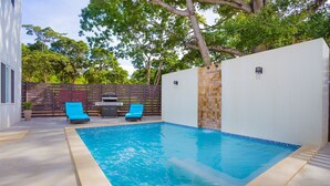 A rectangular swimming pool with blue tiles, surrounded by a white wall and a wooden fence. There are two blue lounge chairs and a barbecue grill against the fence. - New modern condo 3 minute walk to West Bay Beach (West Bay)