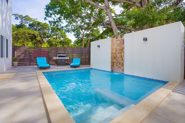Ein rechteckiger Pool mit blauen Fliesen, umgeben von einer weißen Mauer und einem Holzzaun. Gegenüber dem Zaun befinden sich zwei blaue Liegestühle und ein Grill.