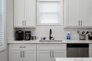A kitchen with white cabinets, a coffee maker, a sink, and a dishwasher. - Barefoot Bliss - Single Level Steps to the Beach (Corolla)