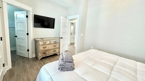 A hotel room with a bed, a wooden dresser, a television mounted on the wall, and a door leading to another room. - Barefoot Bliss - Single Level Steps to the Beach (Corolla)