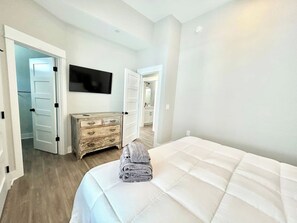 A hotel room with a bed, a wooden dresser, a television mounted on the wall, and a door leading to another room. - Barefoot Bliss - Single Level Steps to the Beach (Corolla)