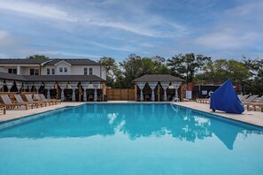 A swimming pool with lounge chairs and covered seating areas, surrounded by buildings and trees. - Barefoot Bliss - Single Level Steps to the Beach (Corolla)