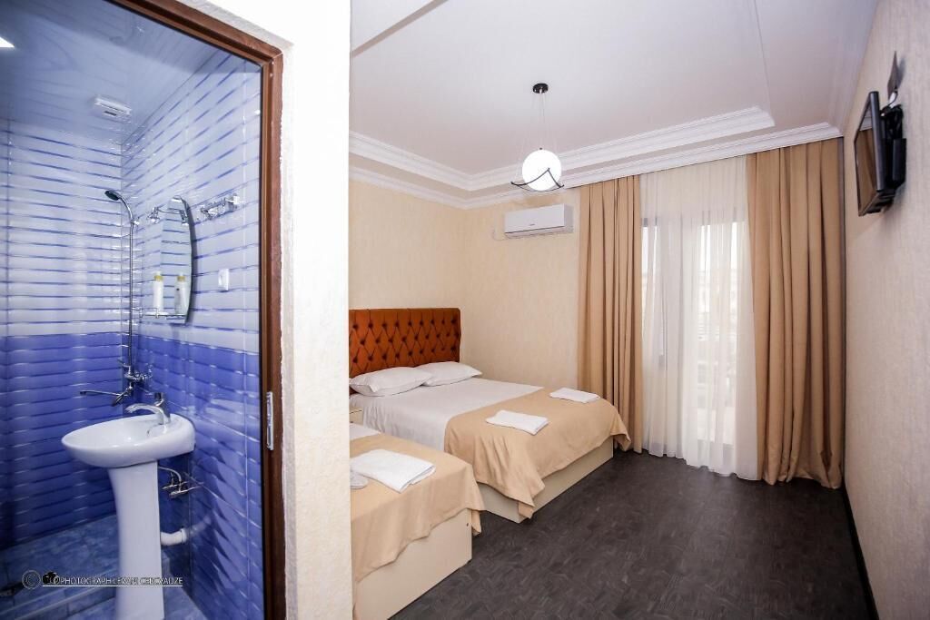 A hotel room with a bathroom featuring a blue-tiled shower, a white sink, and a wooden-framed mirror. The room has two beds with beige bedding, a wooden headboard, and a flat-screen TV mounted on the wall.