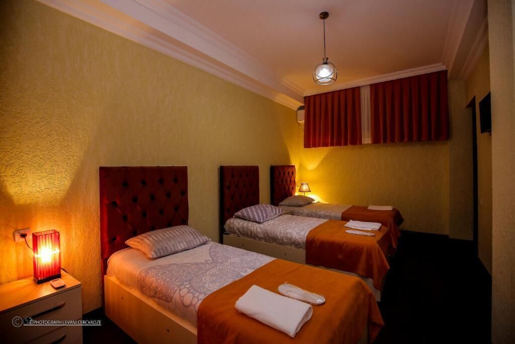 Une chambre d’hôtel avec trois lits, chacun doté d’une tête de lit rouge, d’une table de chevet avec une lampe et d’une fenêtre avec des rideaux rouges.