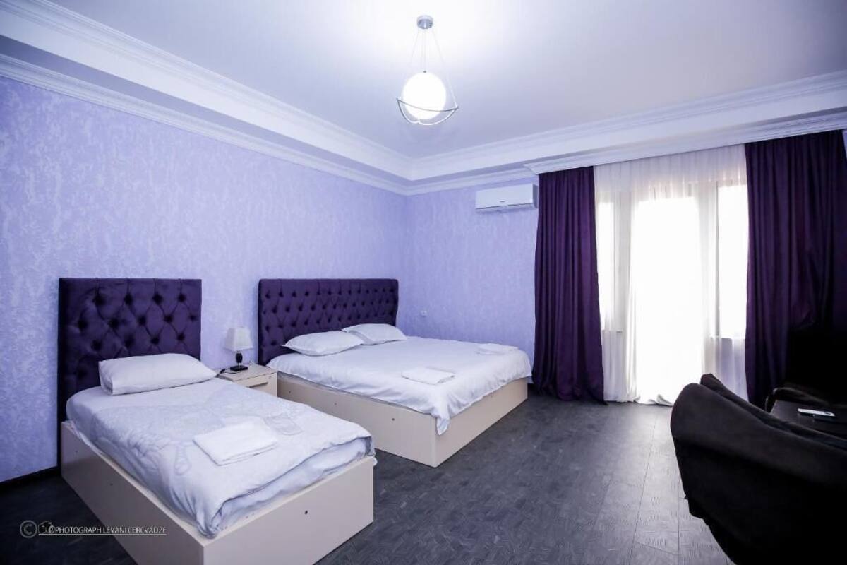 A hotel room with two beds, a large window with purple curtains, a ceiling light, and a chair.