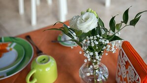 Une table à manger recouverte d’une nappe jaune, avec des chaises blanches et un vase contenant des fleurs et des feuilles vertes.