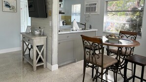 A compact kitchen with a dining area, a TV mounted on the wall, and a window with blinds.
