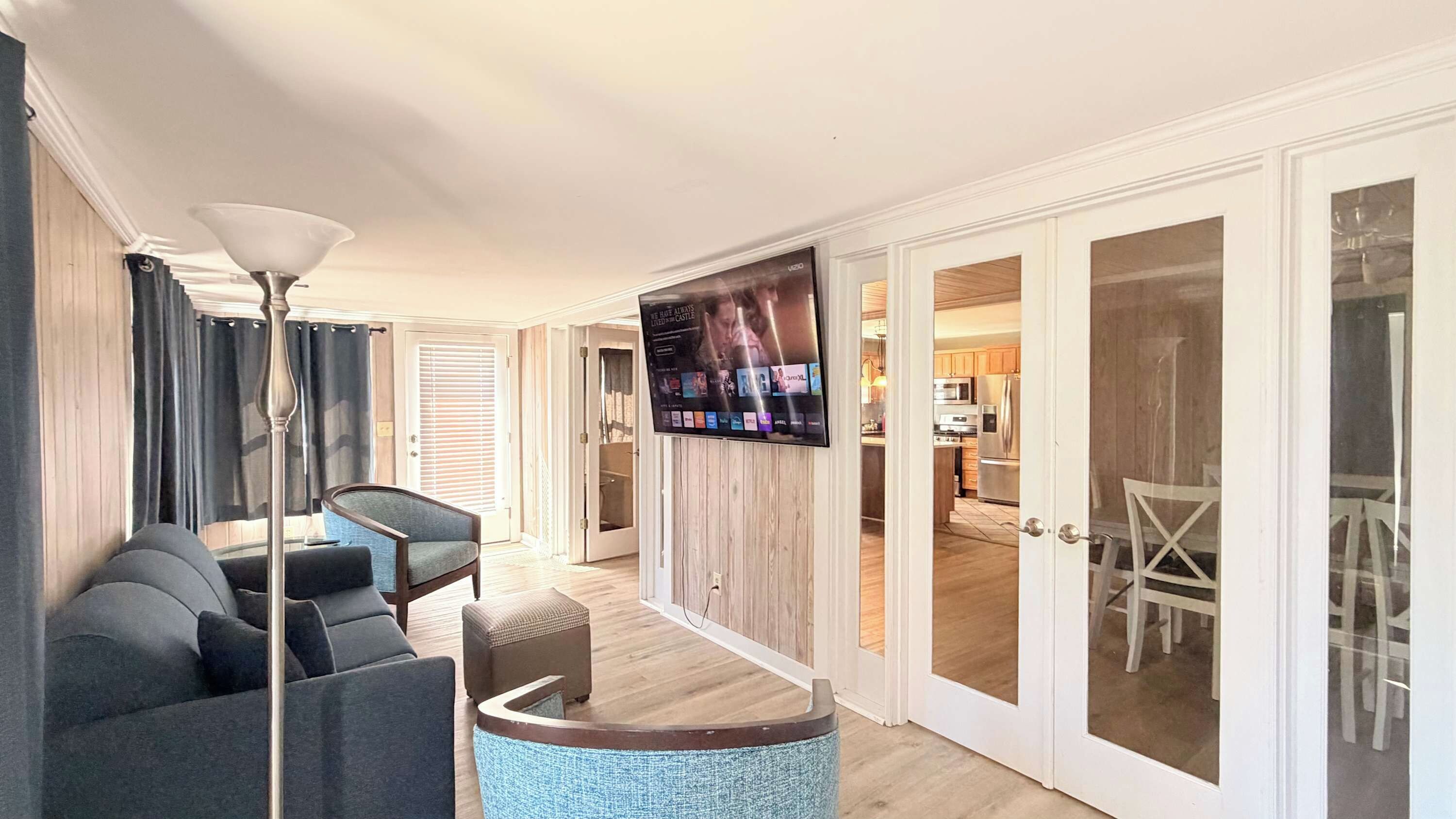 Een moderne woonkamer met een bank, fauteuil en salontafel, een flatscreen televisie aan de muur en een schuifdeur naar een keuken.