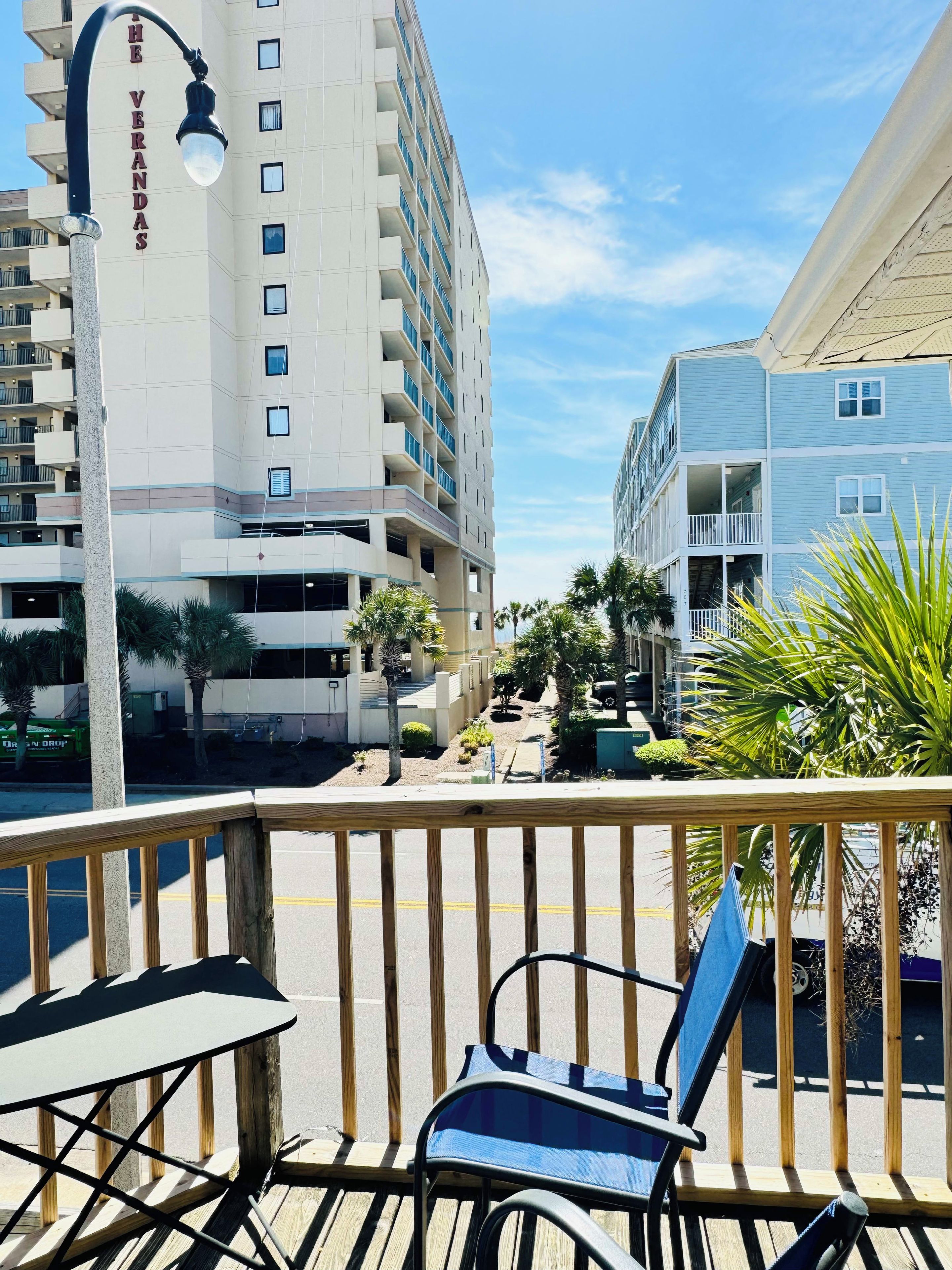A balcony with a chair and table, overlooking a street with buildings and palm trees.