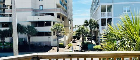 A balcony with a chair and table, overlooking a street with buildings and palm trees.