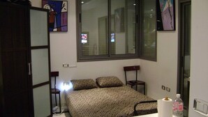 A small, compact bedroom with a bed, a desk, and a chair. There are two windows, one with blinds and one without. - Luxury Studio Apartment - Sea View In Sousse (Port El Kantaoui)