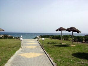A paved pathway leading to the beach with thatched umbrellas and white spherical lights. - Luxury Studio Apartment - Sea View In Sousse (Port El Kantaoui)