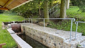 Piscina al aire libre de piedra con escalones de acceso, rodeada de vegetación y una estructura de madera en la parte superior.