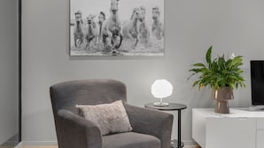 A modern living room with a grey armchair, a small round table with a lamp, a potted plant, and a large black and white horse painting on the wall.