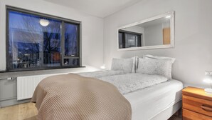 A modern bedroom with a large window, a bed with patterned bedding, a bedside table with a lamp, and a mirror on the wall.