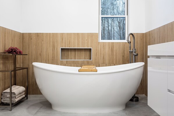 A modern bathroom with a freestanding bathtub, wooden paneling, a window with a view, and a shelf with towels.