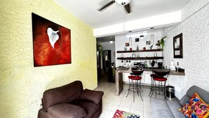 Ein modernes Wohnzimmer mit einem braunen Sofa, einem grauen Sofa mit buntem Kissen sowie einer Küchenzeile mit Barhocker und hängenden Pflanzen.