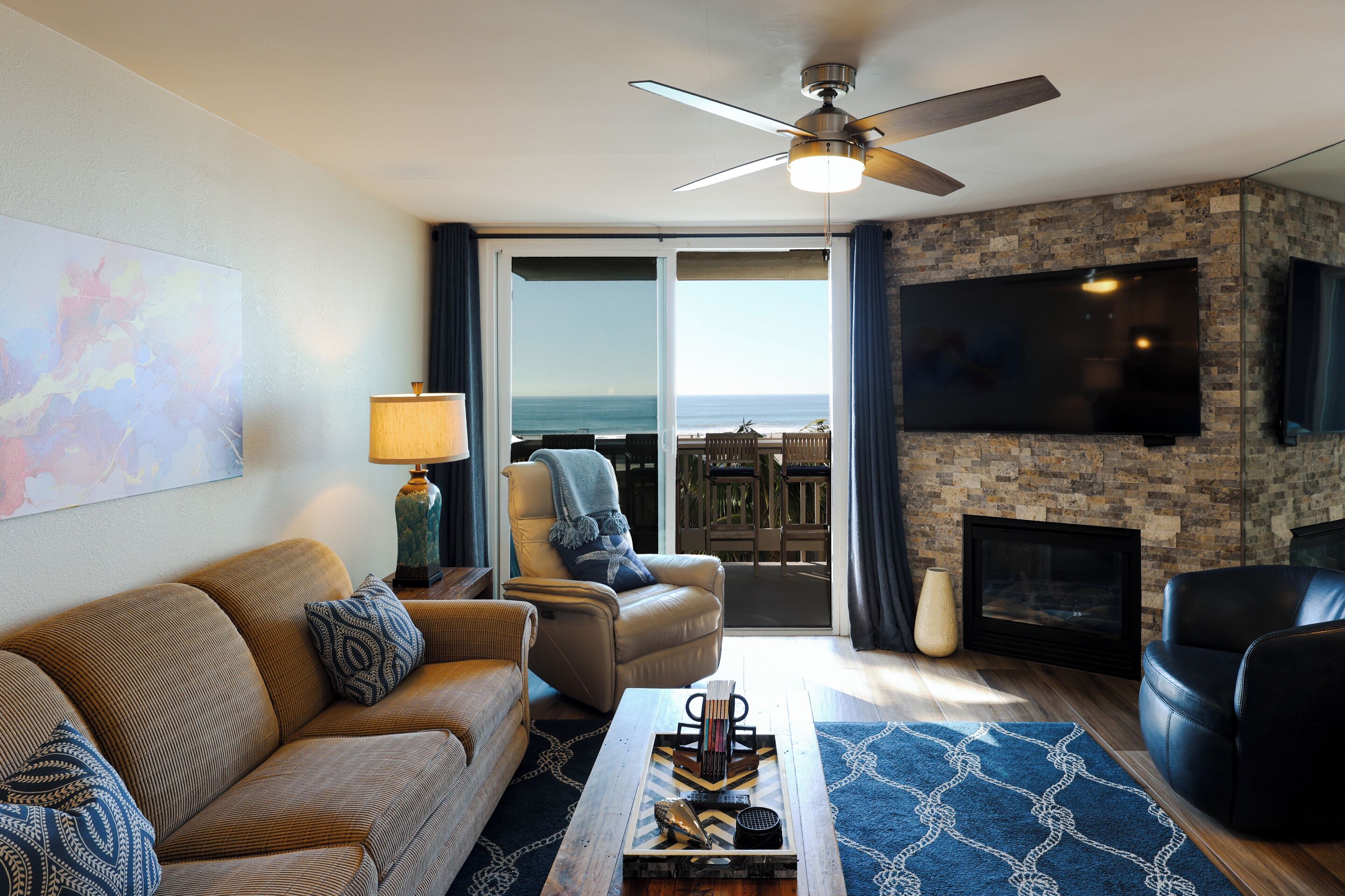 A living room with a sofa, armchair, coffee table, and a fireplace. There is a television mounted on the wall and a ceiling fan.