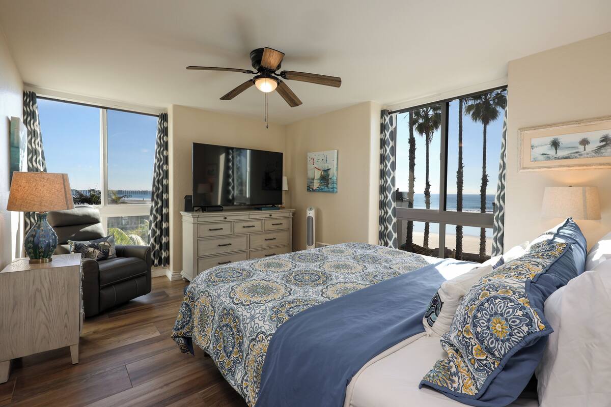 A bedroom with a large bed, a TV, a desk, and a view of the ocean.