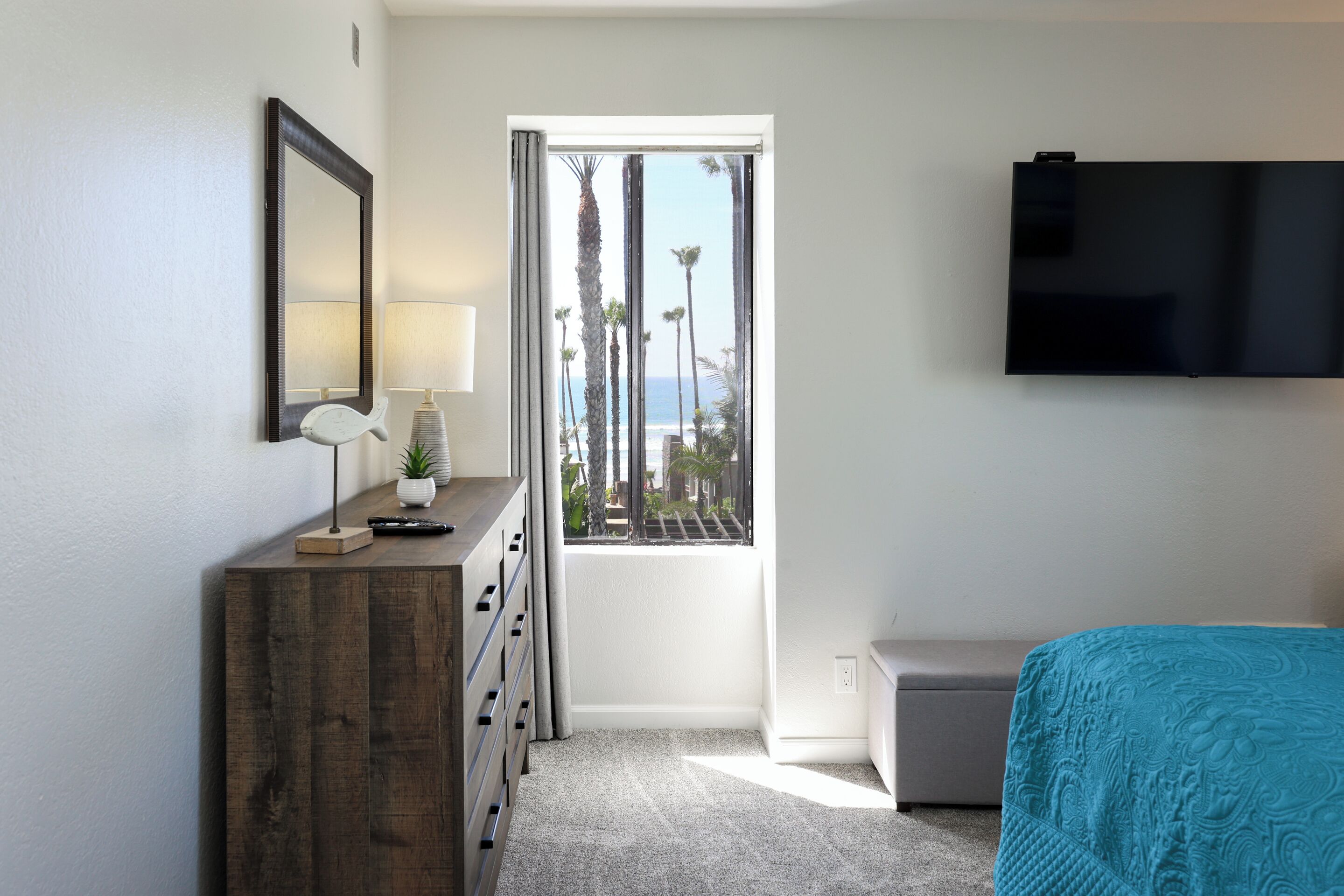 A bedroom with a wooden dresser, a TV mounted on the wall, a window with a view of palm trees and the ocean, and a bed with a patterned bedspread.