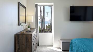 A bedroom with a wooden dresser, a TV mounted on the wall, a window with a view of palm trees and the ocean, and a bed with a patterned bedspread.
