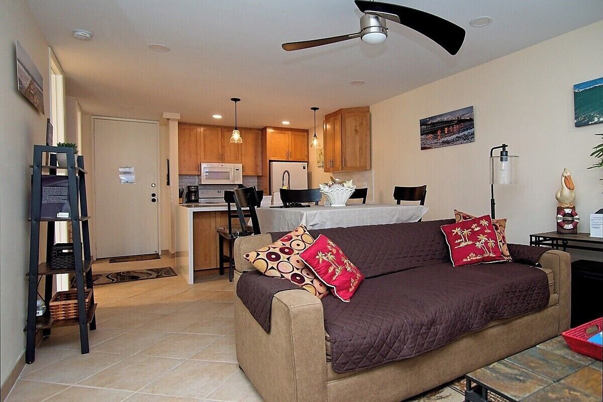 A living room with a sofa, a ceiling fan, and a kitchen in the background.