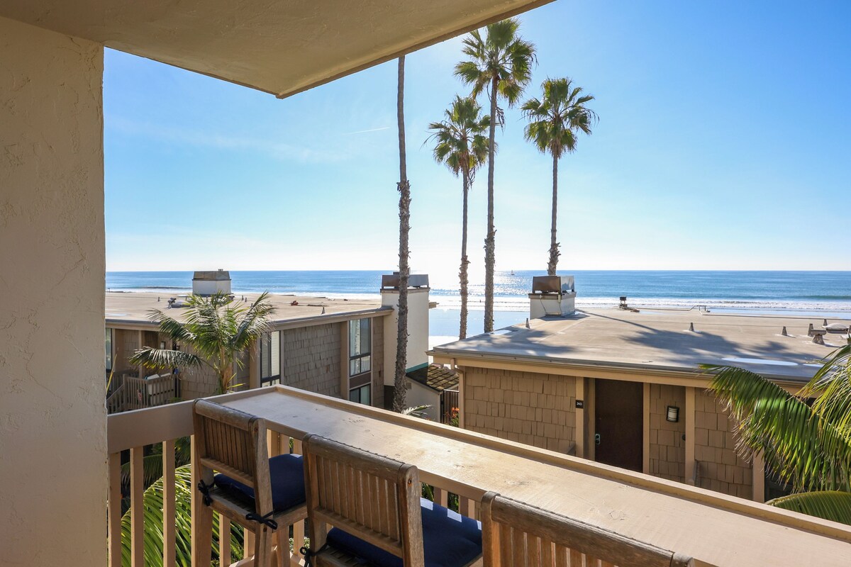 A balcony with a view of the beach, palm trees, and a clear sky.