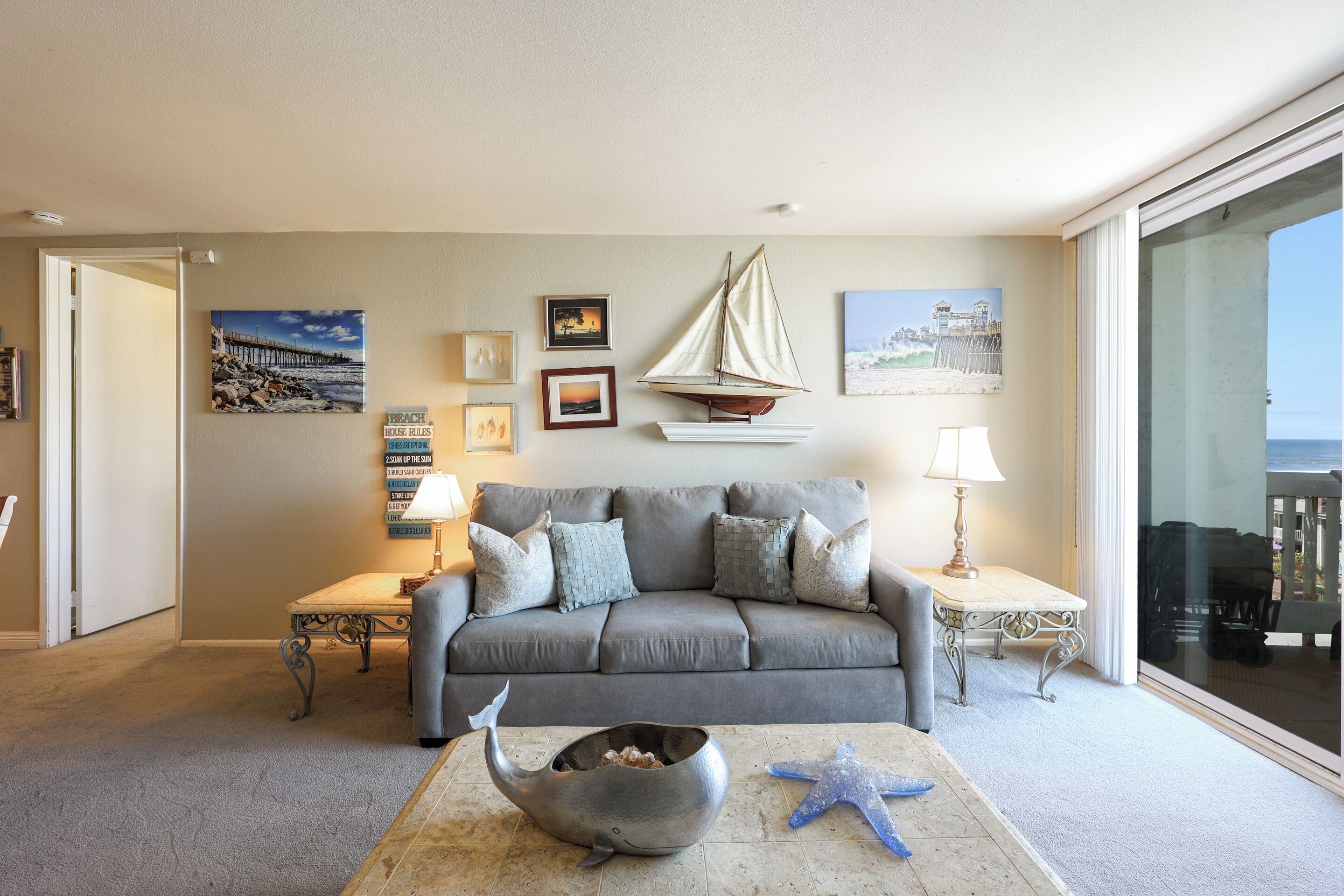 Un salón con un sofá gris, una mesa de centro con una ballena y estrellas de mar, una decoración de velero, y vistas al mar a través de una puerta corredera.