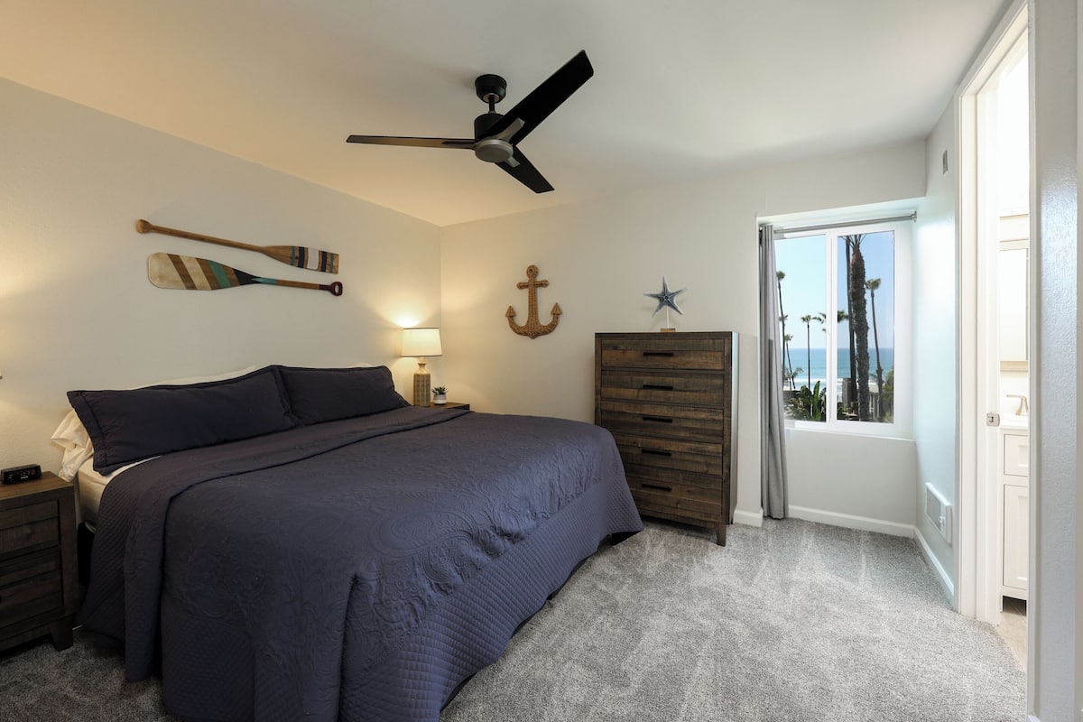 A bedroom with a bed, a ceiling fan, a dresser, a nightstand, and a window with a view.
