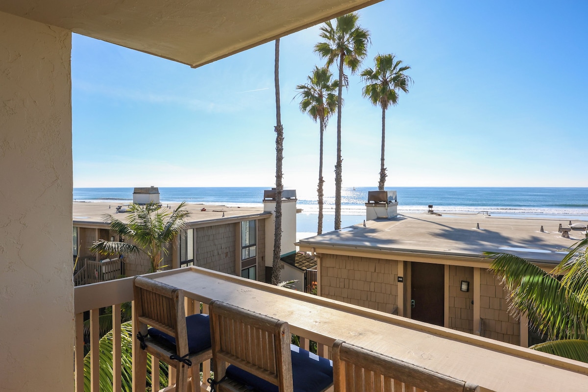 A balcony with a view of the beach, palm trees, and a clear sky.