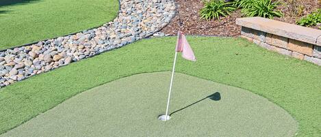 A golf putting green with a flagstick, surrounded by a stone border and a pebble bed.
