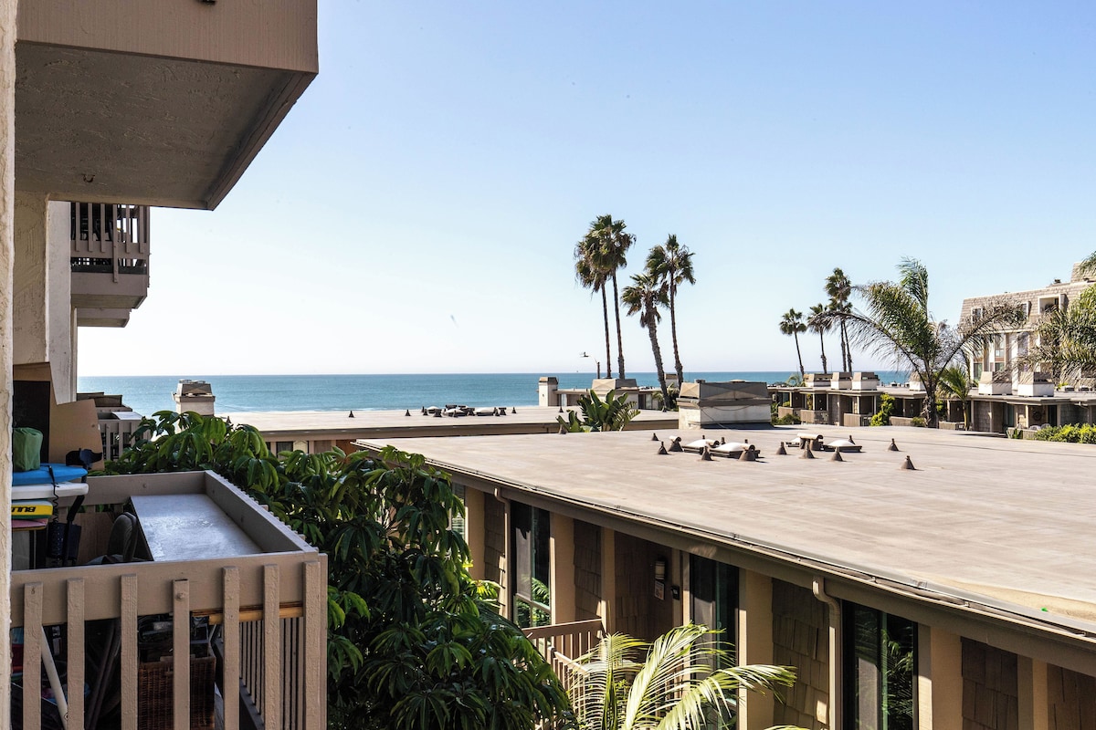 A rooftop terrace with a view of the ocean, palm trees, and a pool area.