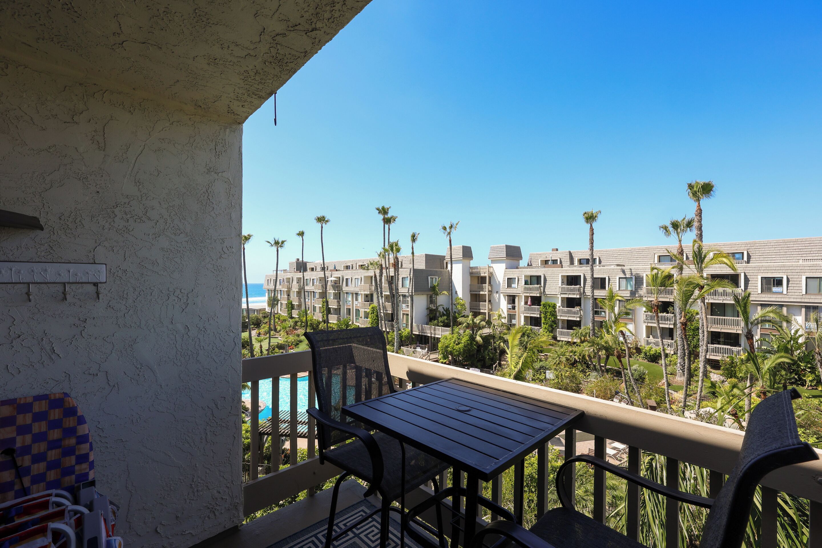 A balcony with a table and chairs, overlooking a pool and a building complex with palm trees.
