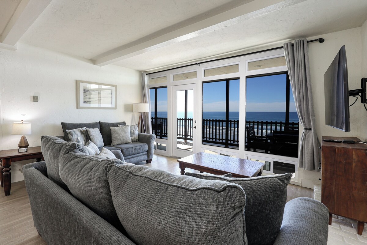 Un salon avec un canapé gris, une table basse et un téléviseur posé sur un meuble en bois. Une grande fenêtre offre une vue sur la mer.