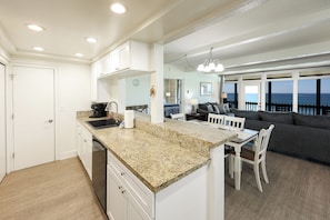 A modern kitchen with granite countertops, white cabinetry, and a dining area with a gray sofa and white chairs. - Beachfront Retreat (Oceanside)