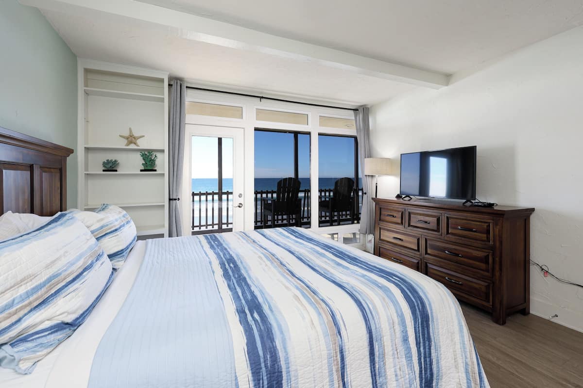 A bedroom with a bed, a dresser, a television, and a view of the sea through a sliding door.
