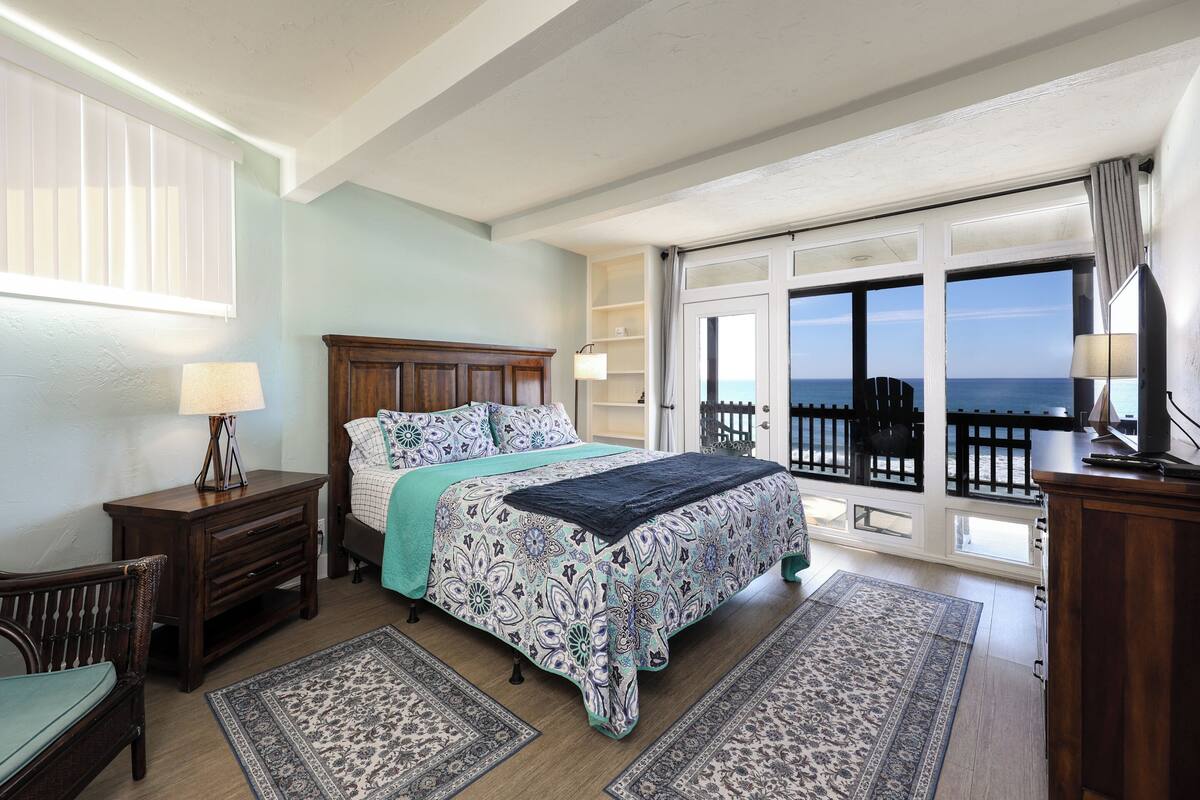 A bedroom with a bed, two nightstands, a chair, a large window with a view of the sea, and a balcony.
