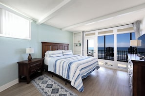 A bedroom with a large bed, a nightstand, a dresser, a window, and a view of the ocean through a sliding glass door. - Beachfront Retreat (Oceanside)