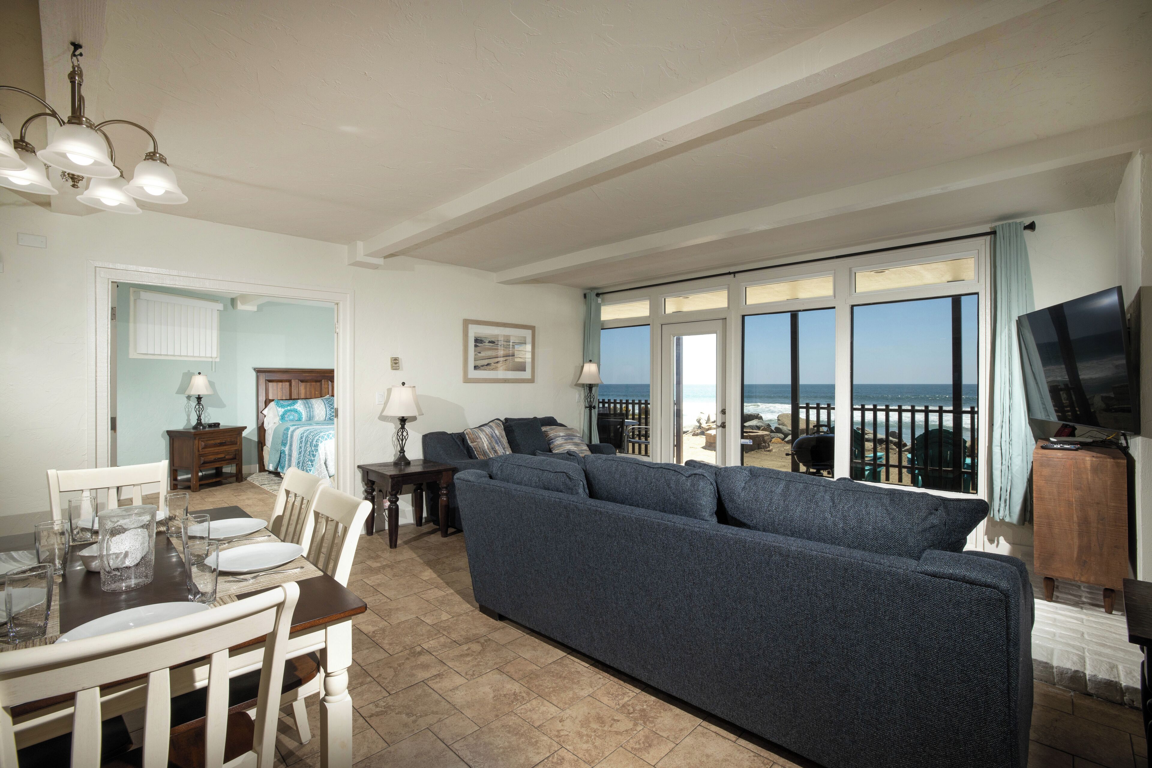 A living room with a large sofa, dining table, and chairs. There is a TV on a wooden stand and a window with a view of the ocean.