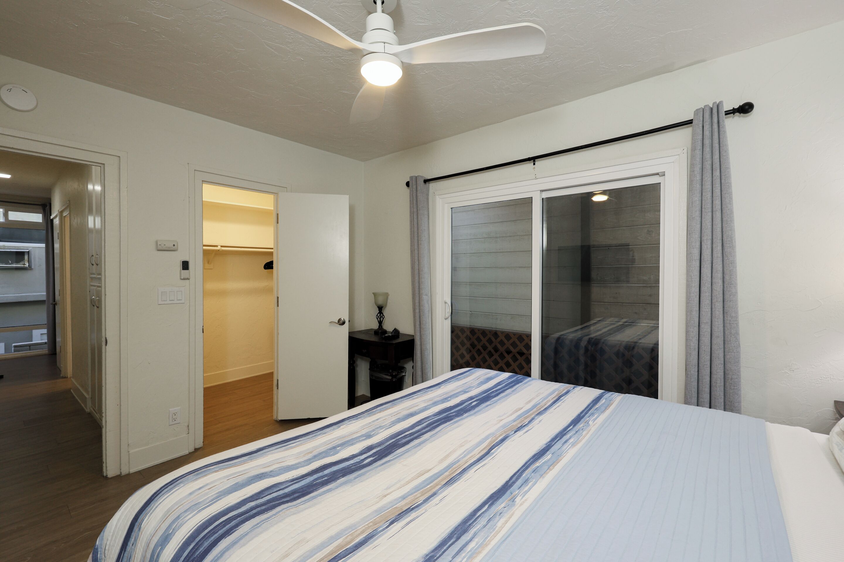 A bedroom with a bed, a ceiling fan, a sliding glass door, and a small table with a lamp.